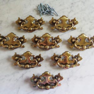 Puede incluir: Conjunto de siete tiradores de cajón antiguos de latón con detalles ornamentados, dispuestos sobre una superficie de mármol. Incluye un pequeño montón de tornillos plateados. Cada tirador presenta un diseño curvo con un asa central y detalles decorativos.