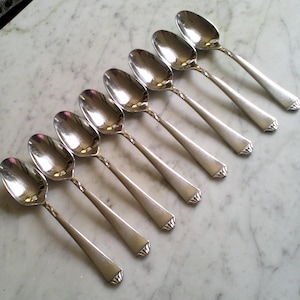 May include: An overhead shot of eight silver spoons arranged diagonally on a marble surface. The spoons have oval bowls and long, tapered handles with a decorative design at the end. The spoons are reflective, showing the light and the marble surface.
