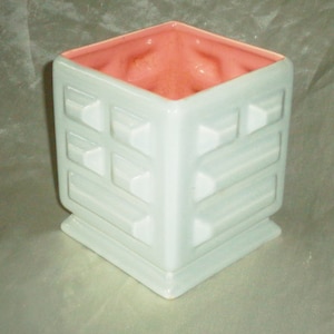 Könnte beinhalten: Ein weißer Keramik-Pflanztopf mit geometrischem Design und rosafarbenem Innenraum. Der Topf hat eine quadratische Form und einen erhöhten Boden.