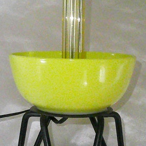 May include: A yellow ceramic lamp base with a black metal tripod stand. The lamp has a silver metal socket and a clear glass shade.