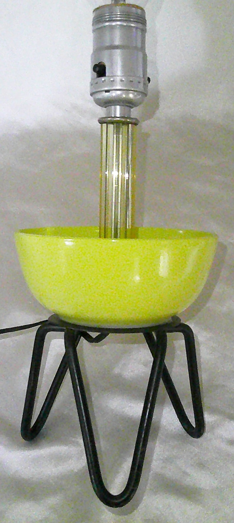 May include: A yellow ceramic lamp base with a black metal tripod stand. The lamp has a silver metal socket and a clear glass shade.