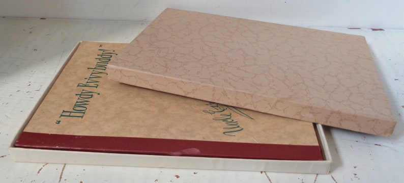 May include: A closed book with a red spine and a white slipcase. The book cover is brown with the text "Howdy Ev'rybuddy!" printed in green. The slipcase is also brown with a textured pattern.