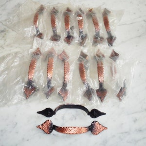 Könnte beinhalten: Ein Set mit 15 kupferfarbenen Schranktürgriffen mit gehämmerter Oberfläche. Die Griffe haben oben und unten eine spitze Pfeilform. Die Griffe sind in durchsichtigen Plastiktüten verpackt.