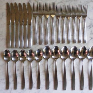 May include: A set of stainless steel flatware, including knives, forks, and spoons, arranged on a marble surface. The silverware has a simple, modern design, reflecting light.