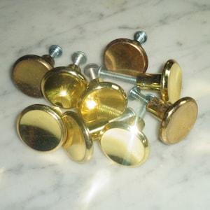 Könnte beinhalten: Eine Gruppe von acht goldfarbenen Schrankknöpfen mit silbernen Schrauben. Die Knöpfe sind rund und haben eine glatte, glänzende Oberfläche.
