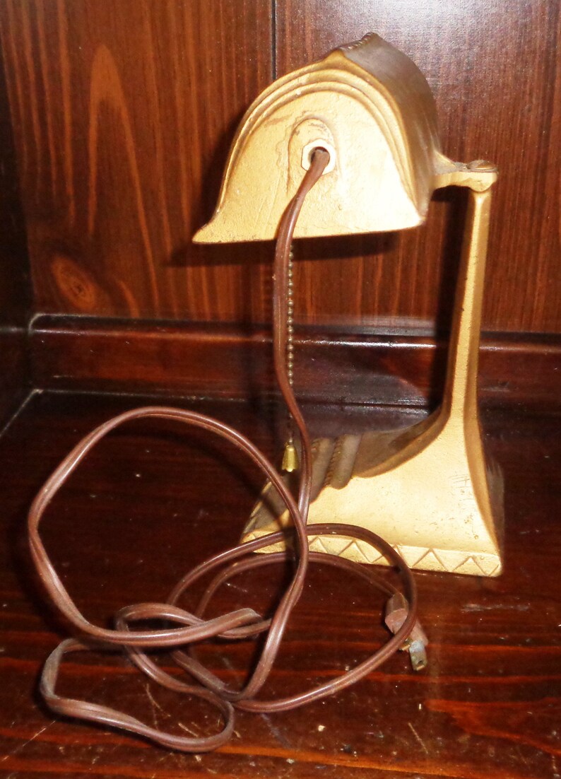 May include: A gold-colored desk lamp with a decorative design. The lamp has a brown cord and a chain hanging from the top.