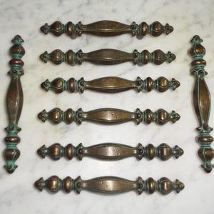 May include: A set of antique bronze cabinet pulls and handles. The pulls have a central oval shape with decorative ends and a verdigris finish. The handles are long with a similar design. The hardware is arranged on a white marble surface.
