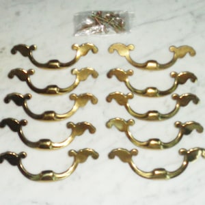 May include: A set of ten brass-colored metal drawer pulls arranged in two columns of five, each featuring a curved, ornate design with decorative ends. A small clear plastic bag containing matching metal mounting screws is positioned at the top.