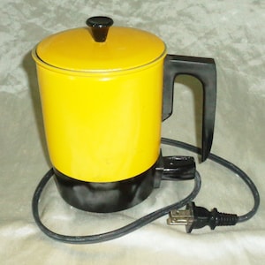 Puede incluir: Una tetera eléctrica amarilla con asa y tapa negras. La tetera tiene una base negra y un cable con enchufe.