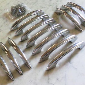 Puede incluir: Un conjunto de tiradores y jaladeras cromadas para armarios, dispuestos sobre una superficie de mármol blanco. Los tiradores varían en tamaño y forma, algunos son largos y curvos, y otros más cortos y angulares. También hay una pequeña bolsa de tornillos.