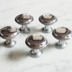 May include: Five silver cabinet knobs with a round, slightly domed top and a raised rim. The knobs have a smooth, shiny finish.
