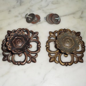 May include: Two ornate, gold-toned cabinet knobs with a decorative, floral design. Each knob has a round, raised center with a smaller, concentric circle. The knobs are accompanied by two sets of screws and washers.