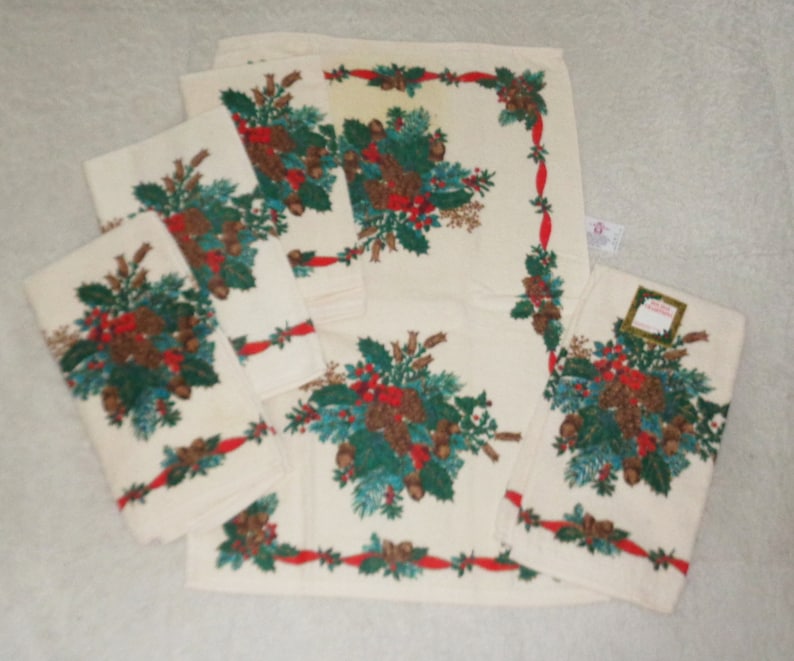 May include: A set of off-white Christmas-themed towels. Each towel features a festive design of pine cones, holly, and red ribbons. The towels are arranged in a neat stack, showcasing the holiday pattern.