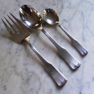 May include: A set of three antique silver utensils: a fork, a large spoon, and a smaller spoon. The fork has four tines, and the spoons have rounded bowls. The utensils are arranged diagonally on a marble surface.