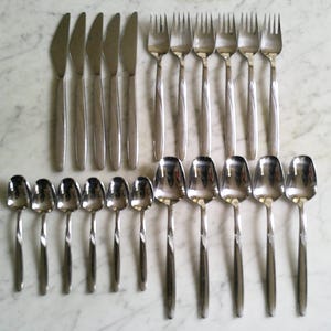 May include: A set of stainless steel flatware consisting of five dinner knives, six forks, six teaspoons, and five tablespoons. Each piece features a smooth, polished finish with a subtle, slender handle design, arranged in rows on a surface.