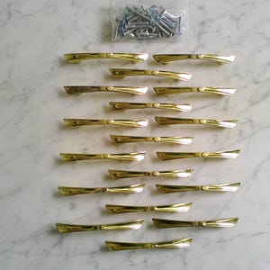 Peut inclure: Ensemble de poignées de meubles dorées disposées sur une surface en marbre. Les poignées ont un design en forme de nœud papillon. Un petit sachet de vis argentées est visible. Quincaillerie pour meubles.