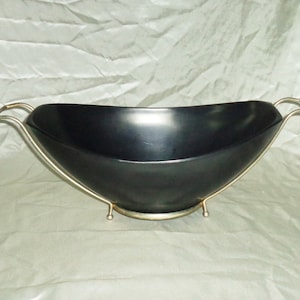 May include: A matte black oval bowl with a smooth interior and exterior, cradled in a gold-toned metal stand. The stand features two side handles and a circular base with small ball feet, elevating the bowl above the surface.