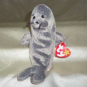 Puede incluir: Foca de peluche gris y blanca con nariz y ojos negros. La foca está sentada sobre una superficie blanca y tiene una etiqueta roja Ty Beanie Baby unida a su aleta derecha.