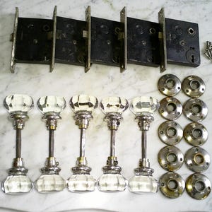 May include: An assortment of antique door hardware, including six glass doorknobs with silver-toned stems, six metal backplates, and several door latches. Also included are screws and other small hardware pieces.
