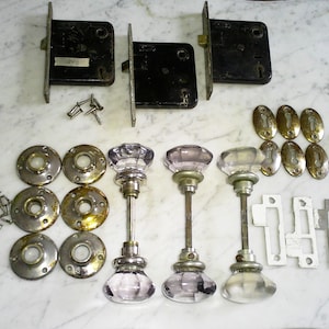 May include: An assortment of vintage door hardware, including three black mortise locks, several brass and silver-toned doorknob rosettes, and three glass doorknobs with silver-toned stems. Also included are brass oval escutcheons and various screws.