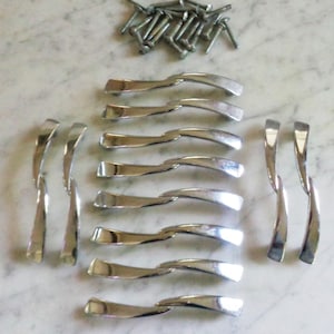 Könnte beinhalten: Eine Sammlung von silberfarbenen Schrankgriffen und Schrauben, die auf einer Marmoroberfläche angeordnet sind. Die Griffe haben ein geschwungenes Design. Die Schrauben sind klein und metallisch.