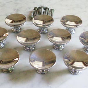 Peut inclure: Un ensemble de dix boutons de meuble argentés avec un design bombé et réfléchissant. Les boutons sont disposés en cercle, avec un groupe de vis visibles en haut. Ces pièces de quincaillerie conviennent à la restauration de meubles.