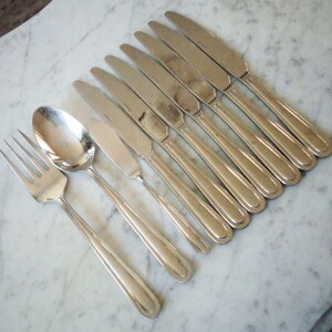 May include: A set of silverware, including a fork, spoon, and eight knives, all made of polished stainless steel. The silverware is arranged in a row on a white marble surface.