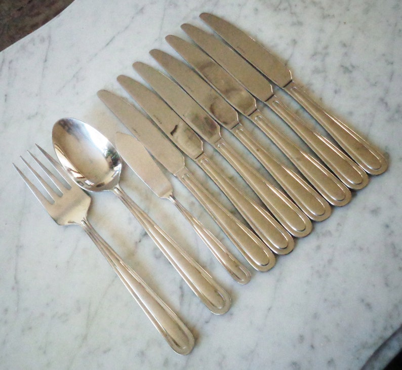 May include: A set of silverware, including a fork, spoon, and eight knives, all made of polished stainless steel. The silverware is arranged in a row on a white marble surface.