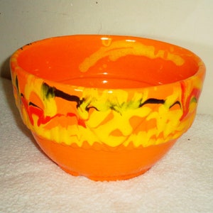 Peut inclure: Bol en céramique orange avec un motif marbré jaune, rouge et noir. Le bol a une forme arrondie et un bord légèrement évasé.