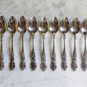 May include: Eight silver spoons with ornate floral detailing on the handles are arranged in a row. The spoons have a reflective, polished surface, and are set against a light-colored, marble-like background. The spoons are likely for dining.