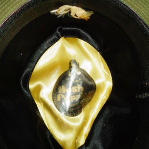 May include: The inside of a black and green hat with a yellow satin lining. A circular label with the text "Hush Puppies" is attached to the lining.