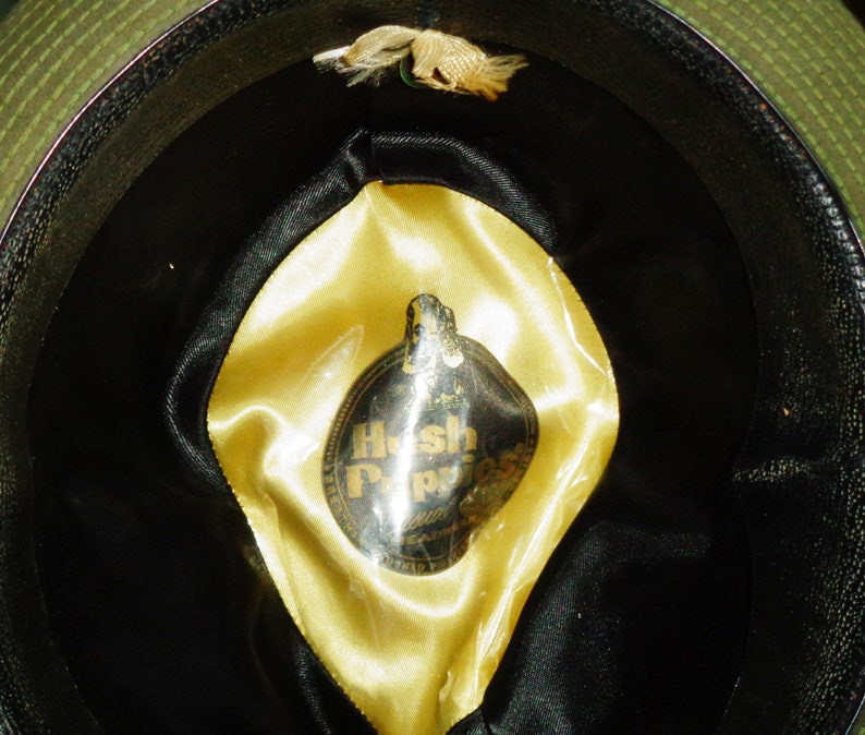 May include: The inside of a black and green hat with a yellow satin lining. A circular label with the text "Hush Puppies" is attached to the lining.