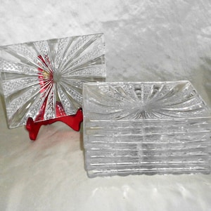 Puede incluir: Una pila de bandejas de servir de vidrio transparente con un patrón de estrella. Las bandejas están apiladas en un soporte rojo.