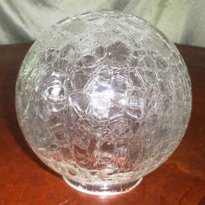May include: A clear, crackle-glass globe with a round shape. The glass has a textured surface with a network of irregular patterns. The globe sits on a small, clear base and is likely a light fixture or decorative element.