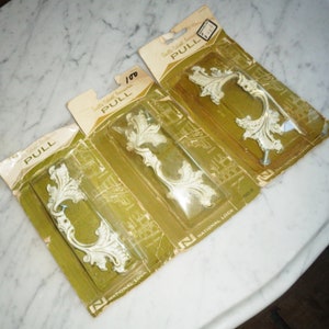 以下が含まれることがあります： 花柄のキャビネットプルが3つ入ったパッケージ。パッケージは黄色で、「Quality Cabinet Hardware」と「PULL」の文字が印刷されています。プルは金属製で、白い仕上げになっています。パッケージには「National Lock」のラベルが貼られています。