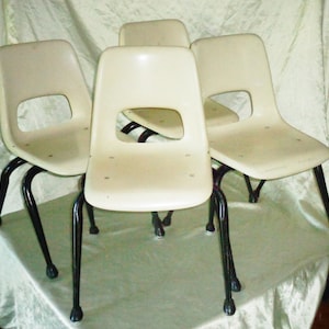 May include: Four cream-colored chairs with black metal legs. Each chair has a curved back with a rectangular cutout and a slightly angled seat. The legs are slender and splayed, providing stability.