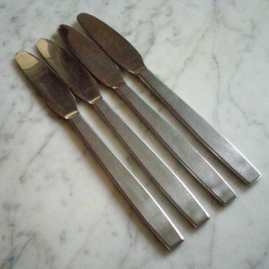 May include: A set of four silver-toned stainless steel dinner knives with a brushed matte finish. Each knife features a long, flat, rectangular handle and a rounded, smooth blade edge, arranged diagonally in a parallel row on a flat surface.