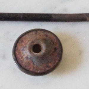 May include: A rusty metal bottle stopper with a round, flat top and a small hole in the center. The stopper is attached to a long, thin metal rod.