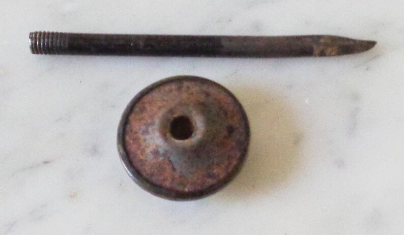 May include: A rusty metal bottle stopper with a round, flat top and a small hole in the center. The stopper is attached to a long, thin metal rod.