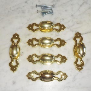 May include: Set of six gold-toned cabinet knobs with an oval-shaped center and decorative scrollwork. The knobs are designed for use on drawers or cabinets.