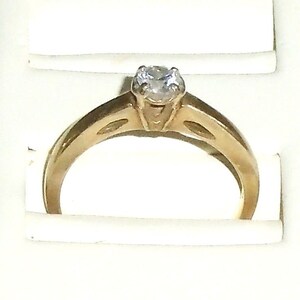 May include: A gold-colored engagement ring with a single, clear gemstone. The ring has a classic design with a raised setting for the stone and a smooth, polished band. The ring is displayed in a white box.