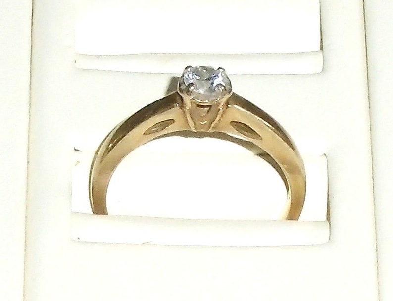 May include: A gold-colored engagement ring with a single, clear gemstone. The ring has a classic design with a raised setting for the stone and a smooth, polished band. The ring is displayed in a white box.