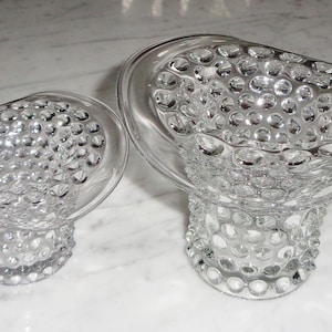 Può includere: Due vasi di vetro trasparente con un designo testurizzato a forma di bolle. Il vaso più grande ha un manico. Entrambi i vasi sono su una superficie di marmo bianco.
