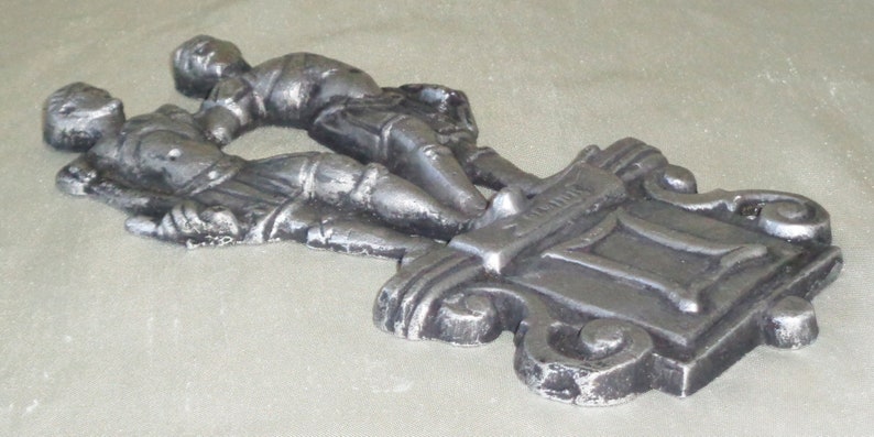 Puede incluir: Una decoraci&oacute;n de pared de metal plateado con dos figuras que se toman de la mano, con la palabra "G&eacute;minis" inscrita en la base.