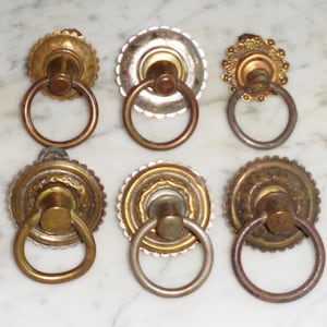Könnte beinhalten: Sechs antike Messing-Schubladengriffe mit verzierten Designs und Ringgriffen. Die Griffe haben verschiedene Oberflächen, darunter poliertes Messing, gealtertes Messing und eine silberfarbene Oberfläche.