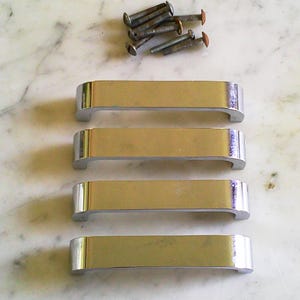 Puede incluir: Cuatro tiradores de armario rectangulares con acabado en cromo pulido, dispuestos en una columna vertical. Cada tirador presenta una superficie lisa y plana con extremos redondeados. Un pequeño grupo de tornillos metálicos está encima.