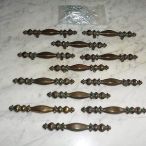 May include: A collection of ten matching metal drawer pulls with a dark bronze finish and a central spindle design. Each handle features decorative detailing along the length and includes a small plastic bag containing mounting screws for installation.