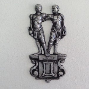 Puede incluir: Una escultura de metal plateado del signo zodiacal G&eacute;minis. Dos figuras se paran una al lado de la otra en un pedestal con la palabra "G&eacute;minis" inscrita en &eacute;l.