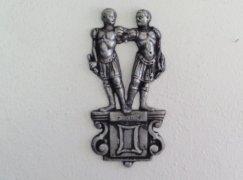 Puede incluir: Una escultura de metal plateado del signo zodiacal G&eacute;minis. Dos figuras se paran una al lado de la otra en un pedestal con la palabra "G&eacute;minis" inscrita en &eacute;l.