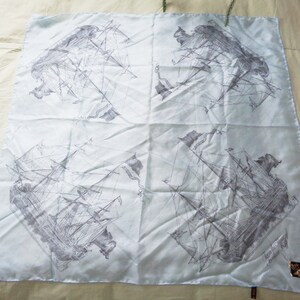 May include: A light blue square scarf featuring a repeating pattern of detailed gray ship illustrations. The scarf has a subtle sheen and a delicate appearance, with a small tag in the corner.